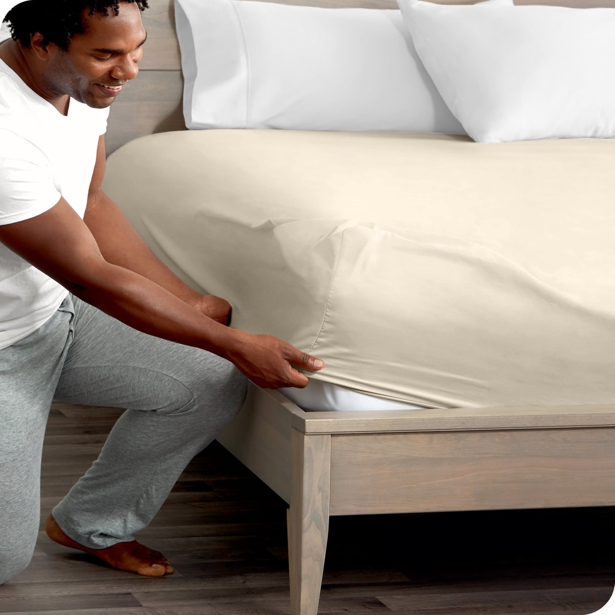 Man in white shirt and gray pants adjusting a fitted sheet on a bed.