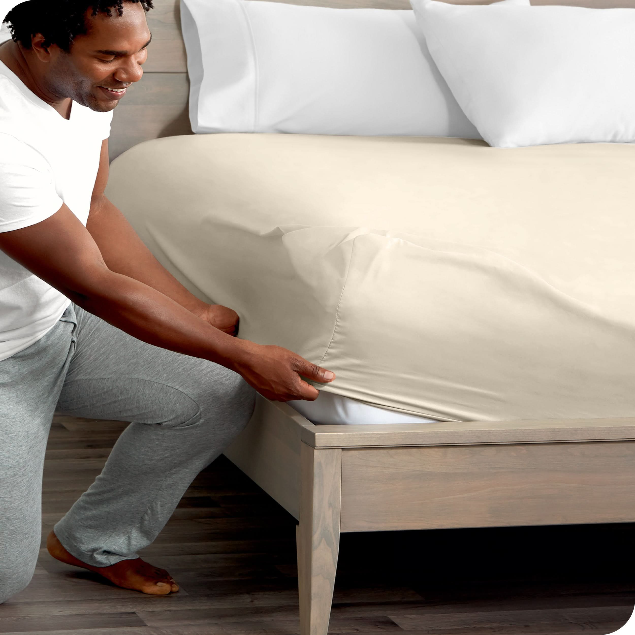 A man is kneeling while putting a fitted sheet on a bed