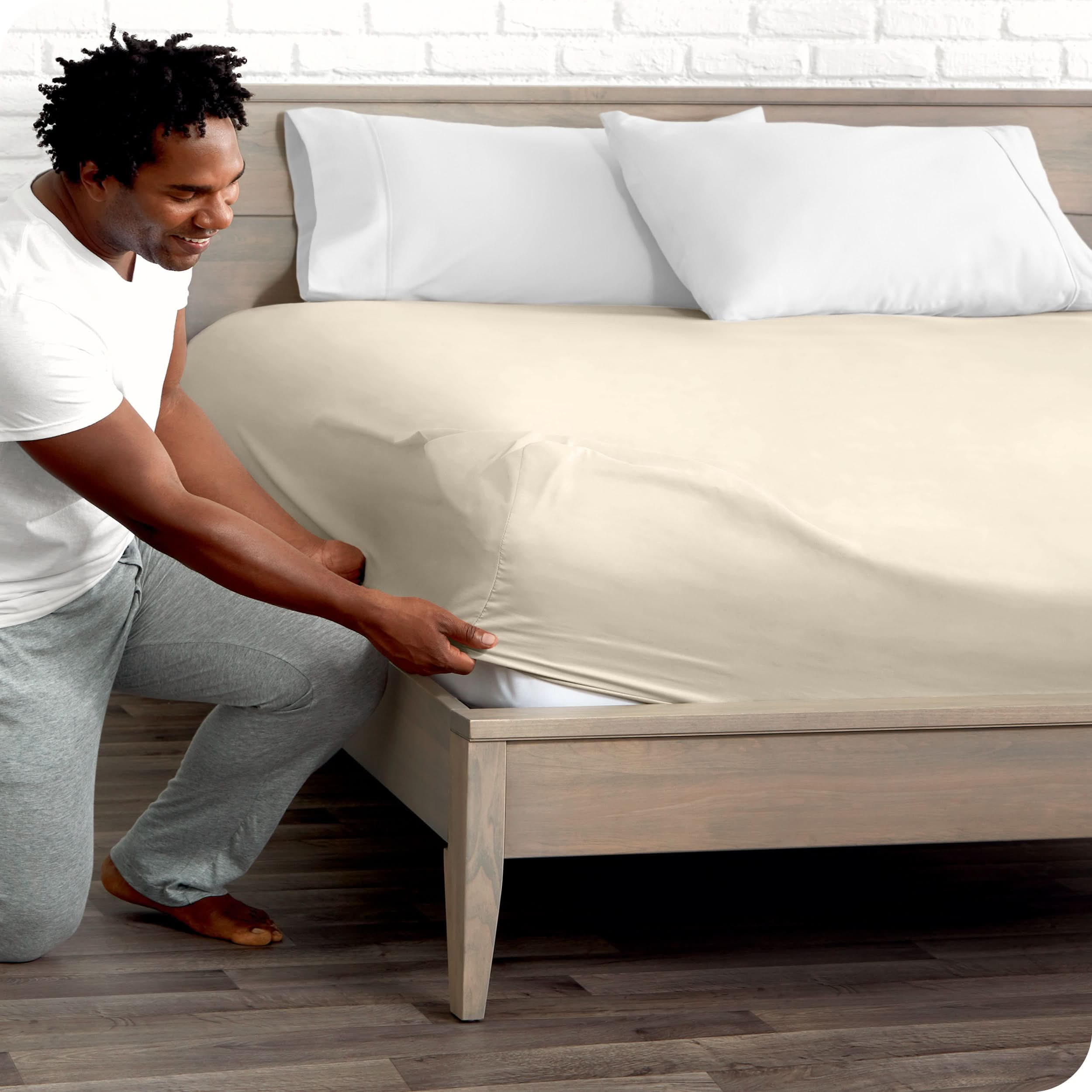 A man is kneeing and putting the corner of the fitted sheet on the mattress. The mattress is on a wooden bed frame.