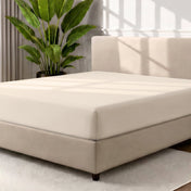 A fitted sheet covering a bed with a cream colored frame, styled in a bedroom setting with a large potted plant.