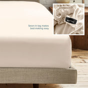A fitted bed sheet on a bed frame with a small inset close-up of the elastic and a sewn-in tag labeled BOTTOM. White text on the sheet reads, 'Sewn-in tag makes bed-making easy.