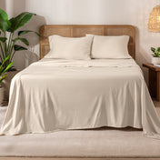 Organic cotton jersey sheets on a bed with a wicker headboard in a room with plants and a lamp.
