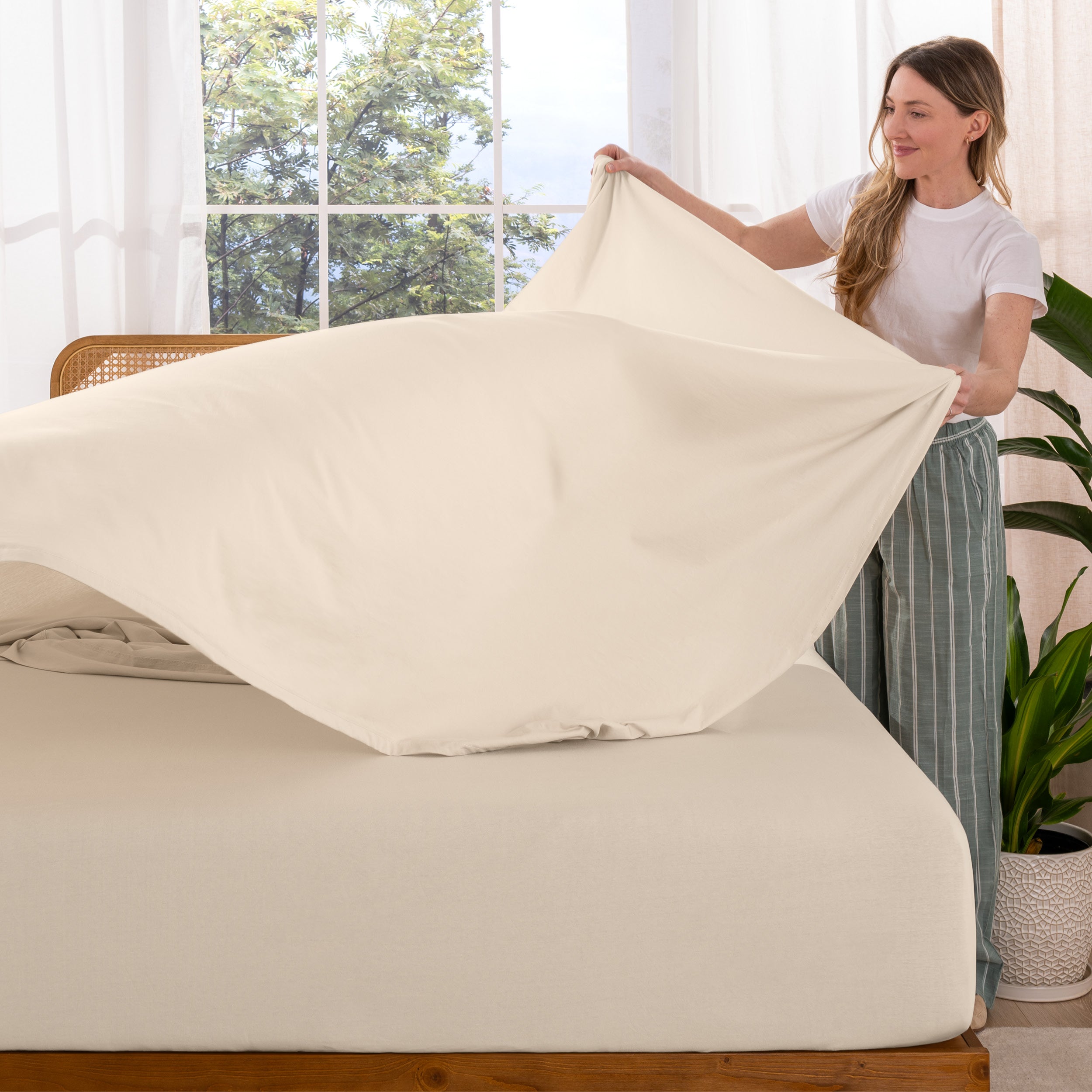 Woman putting sheet on a bed in a bright room with large windows.