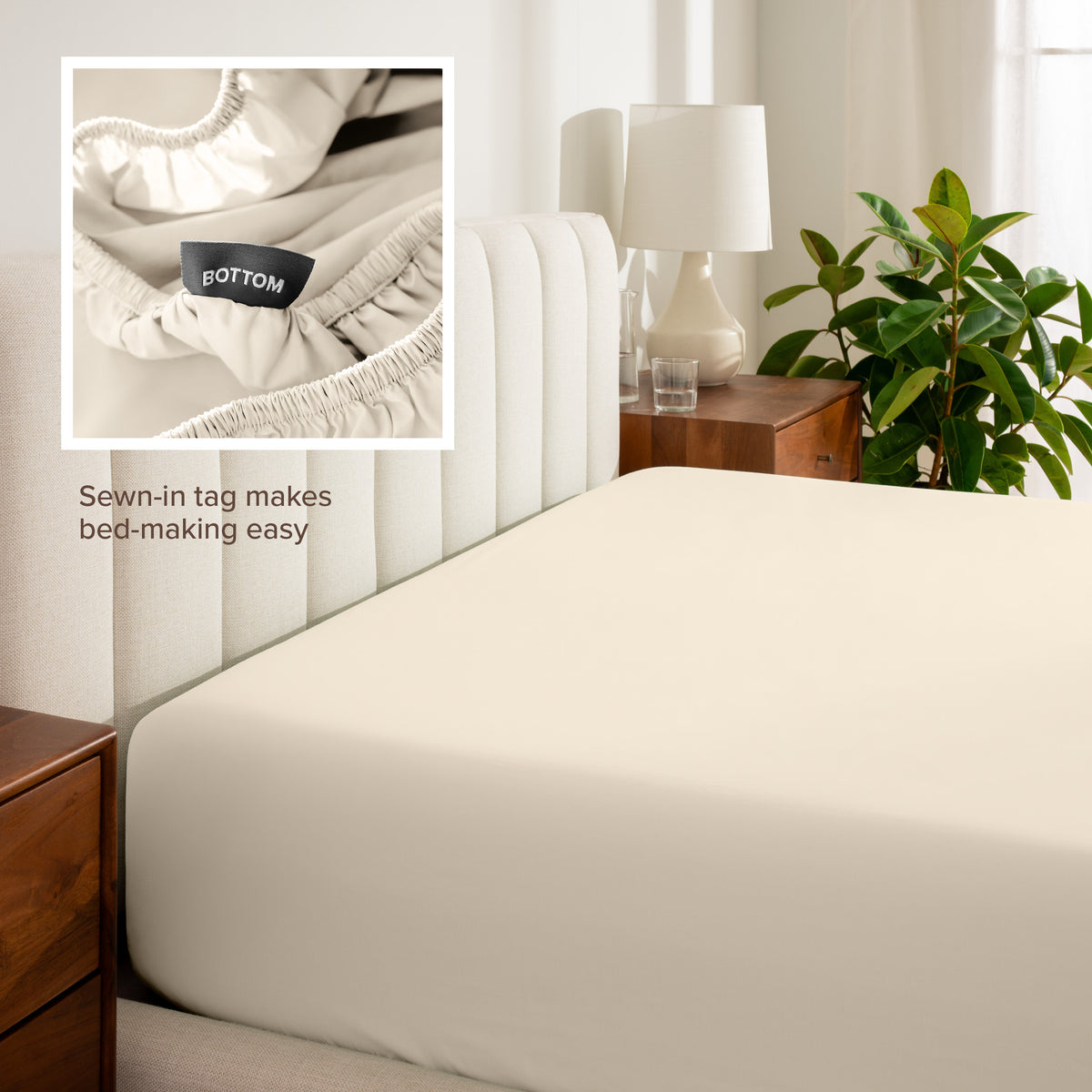Fitted sheet on a bed with a close-up of a sewn-in tag, nightstand, lamp, and plant in a bedroom setting.
