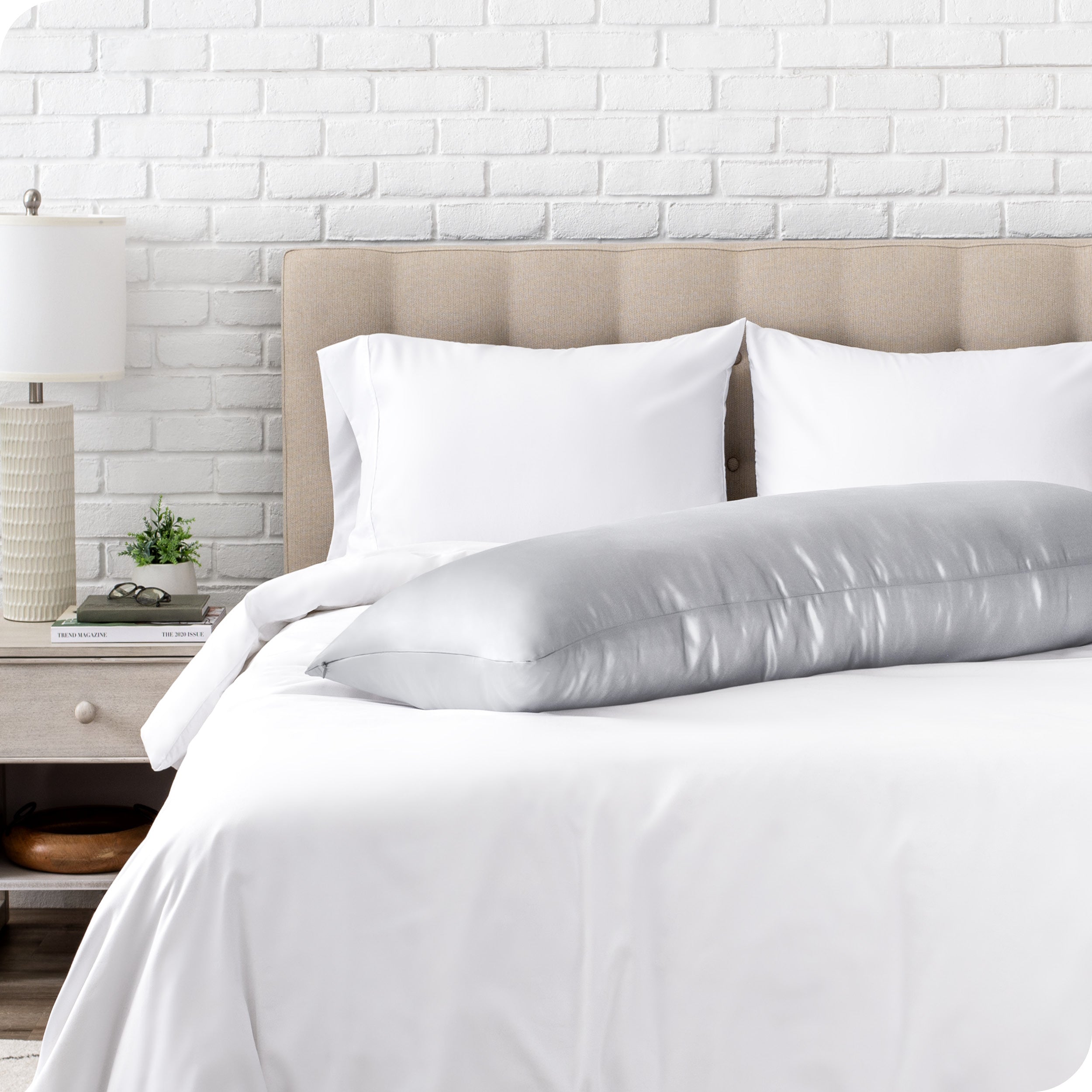 Bed with white bedding and a body pillow against a white brick wall.