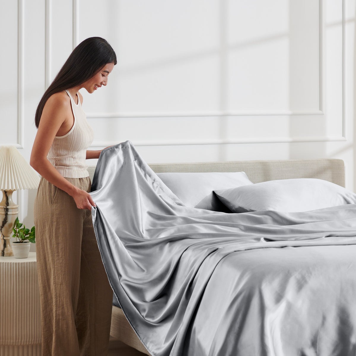 Woman arranging satin bedding on a bed in a bright bedroom.