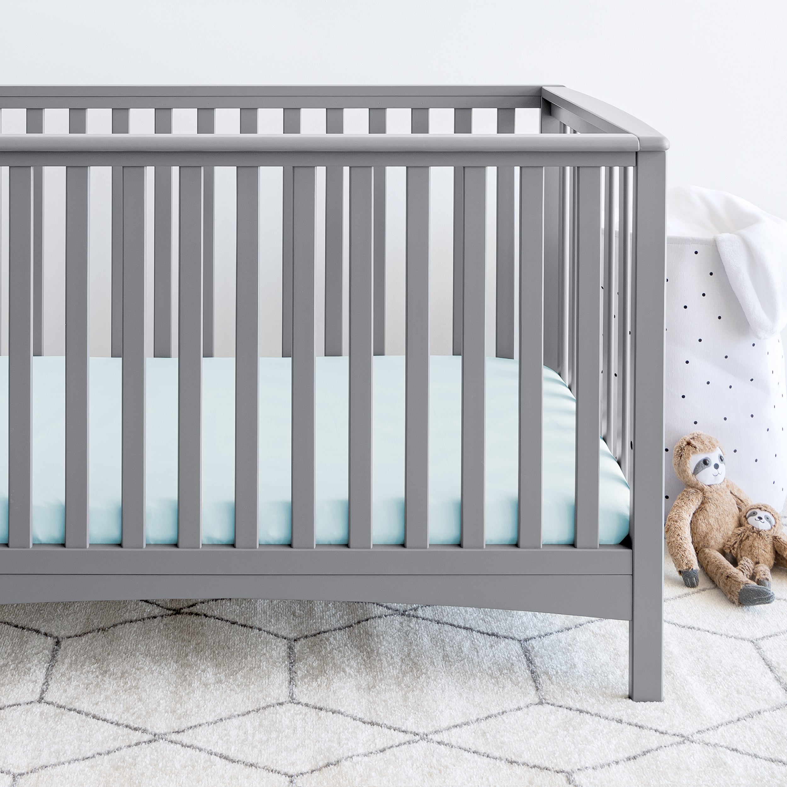 A gray crib on a white printed rug. The crib mattress is cover with a blue fitted sheet. Sloth stuffed animals and a hamper are next to the crib. 