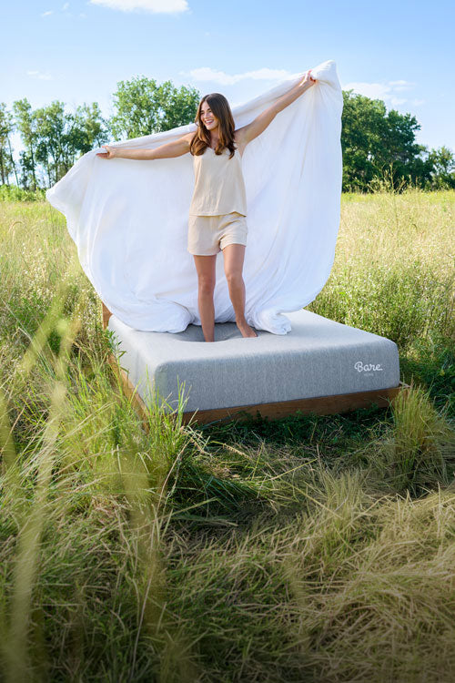 Woman standing on a bare mattress in a grassy field with a white sheet.