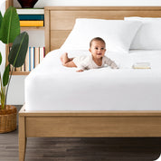 Terry mattress protector on a mattress with 2 pillows on top near the headboard. A baby is lying on the bed next to a bottle.