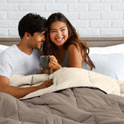Man and woman sitting on a bed holding a mug, smiling at each other.