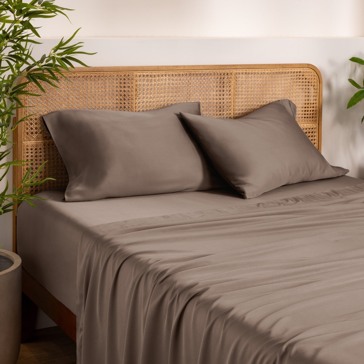 Bamboo bed sheets and pillows on a bed with a wicker headboard in a room with plants.