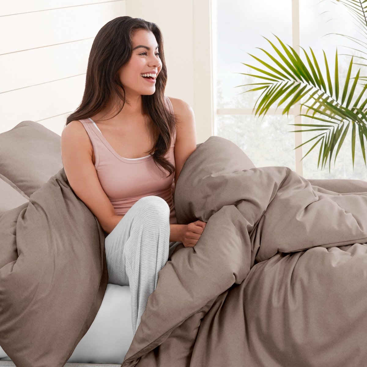 Woman sitting on a bed under duvet cover with a plant in the background