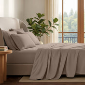 A neatly made bed with in a softly lit bedroom. A large green plant stands beside the bed, and a window with curtains is visible in the background.