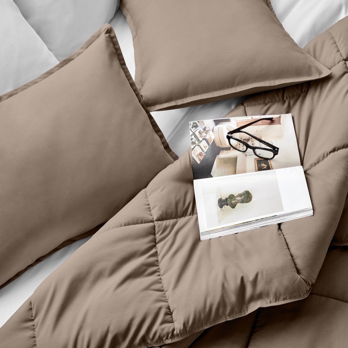 Comforter and pillows on a bed with a magazine and glasses.