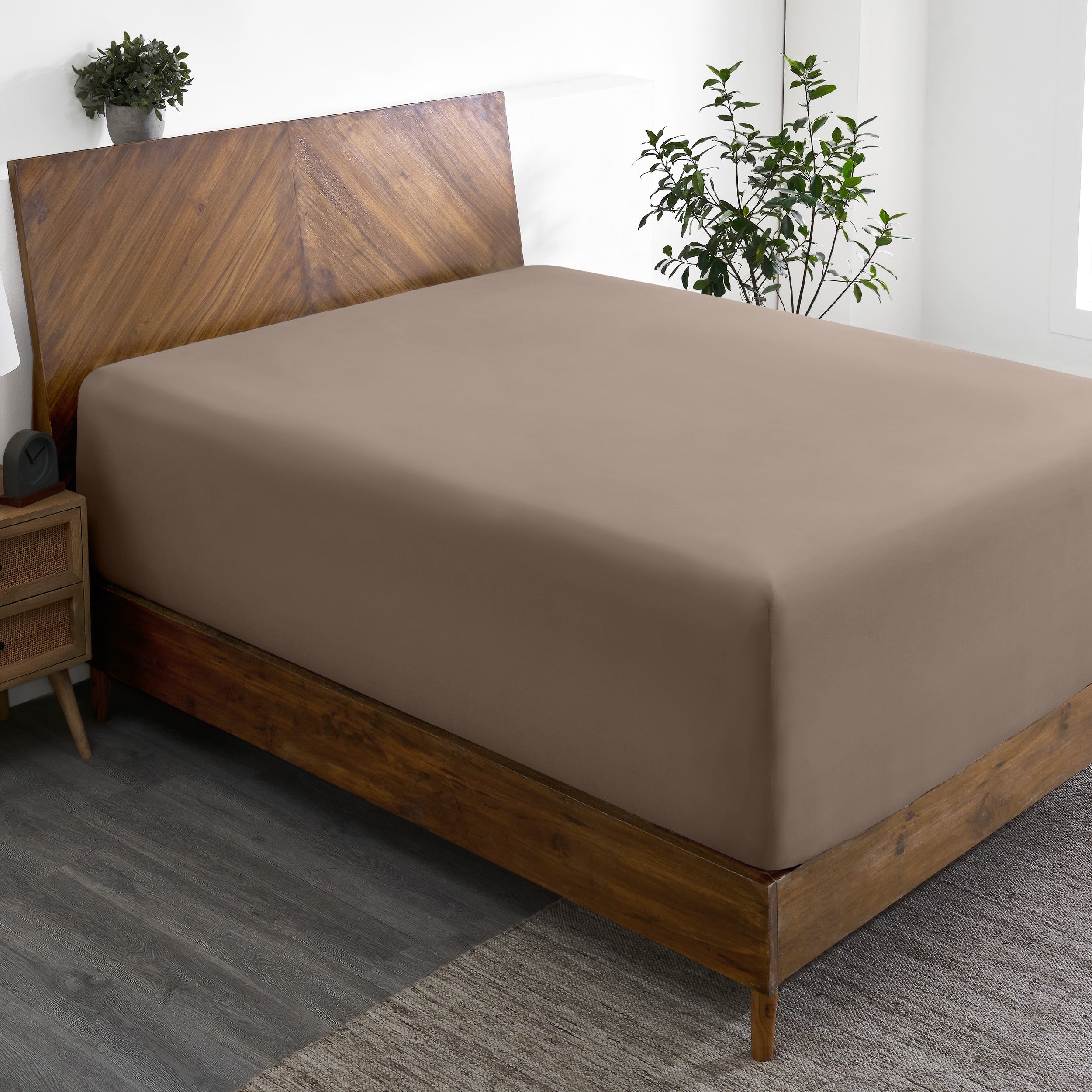 Wooden bed frame with a fitted sheet on the mattress in a room with plants and a nightstand.