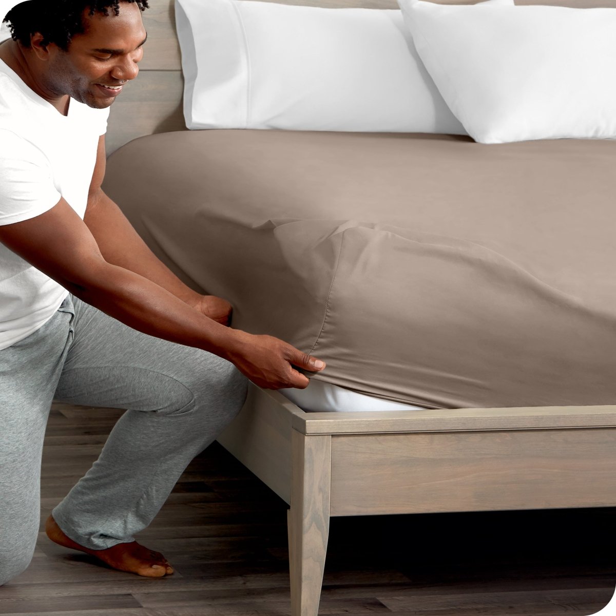 Man in white shirt and gray pants adjusting a fitted sheet on a bed.