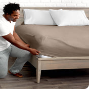 A man is kneeing and putting the corner of the fitted sheet on the mattress. The mattress is on a wooden bed frame.