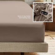 A fitted bed sheet on a bed frame with a small inset close-up of the elastic and a sewn-in tag labeled BOTTOM. White text on the sheet reads, 'Sewn-in tag makes bed-making easy.