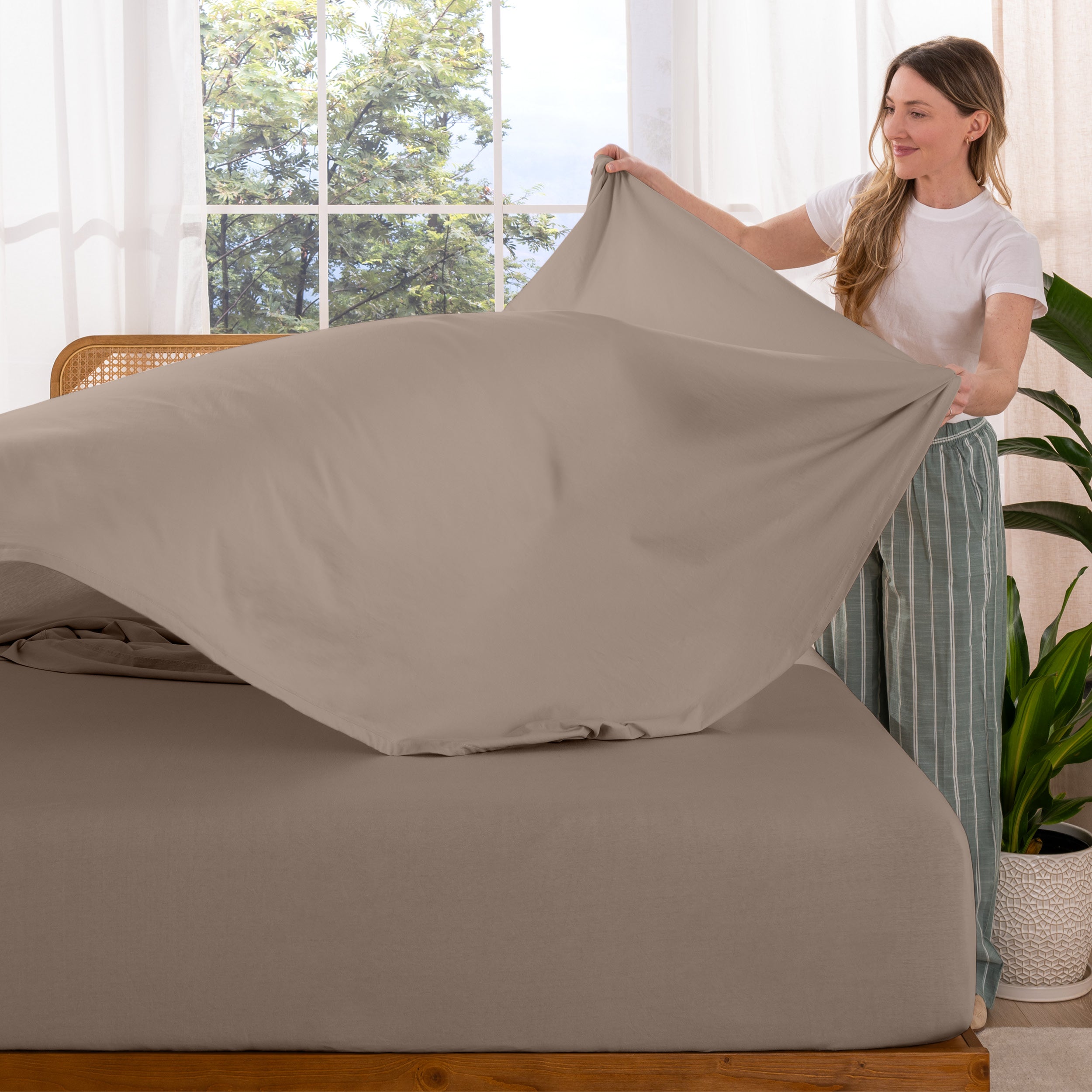 Woman putting sheet on a bed in a bright room with large windows.