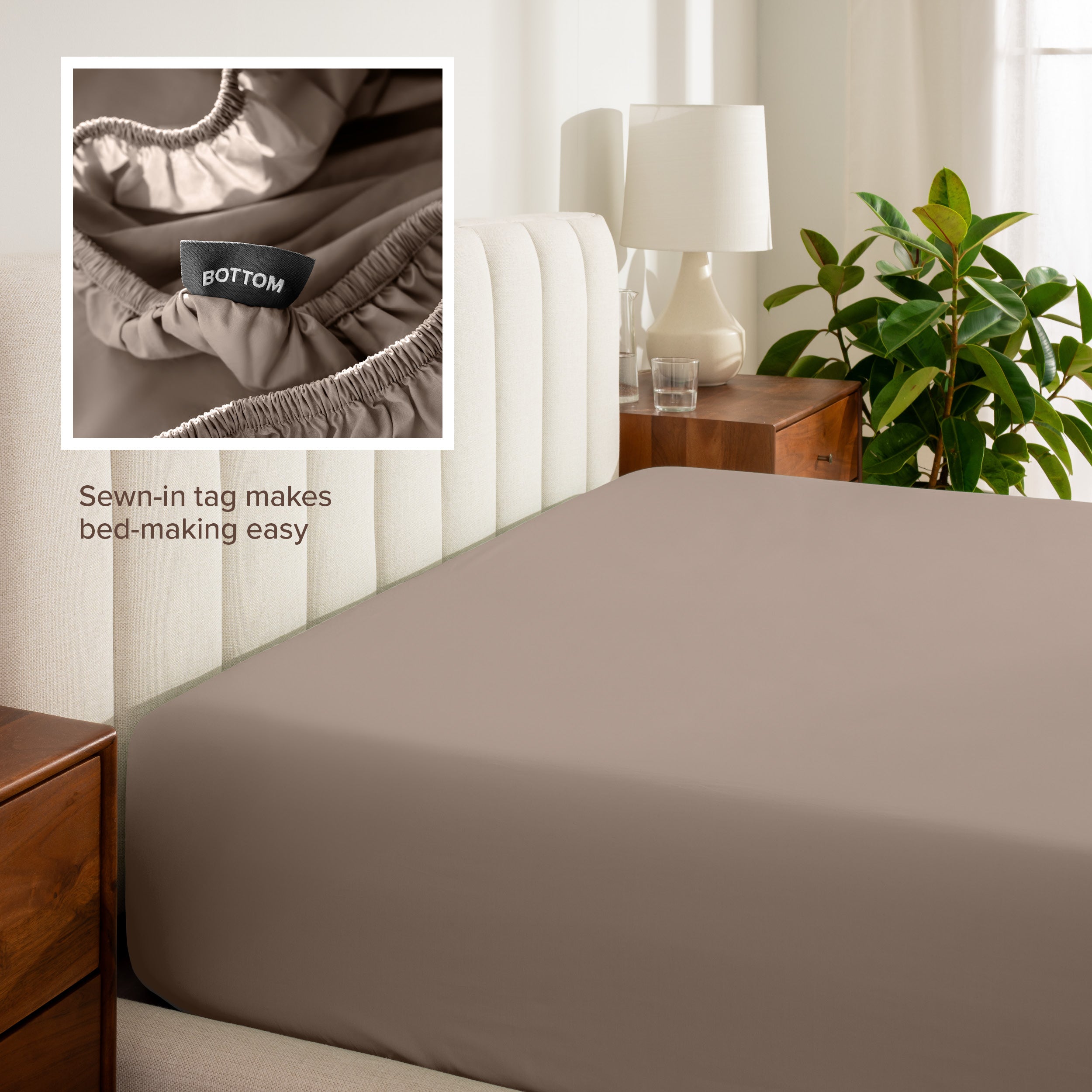 Fitted sheet on a bed with a close-up of a sewn-in tag, nightstand, lamp, and plant in a bedroom setting.