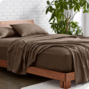 Bed with heather sheets and wooden frame against a white brick wall with a plant.