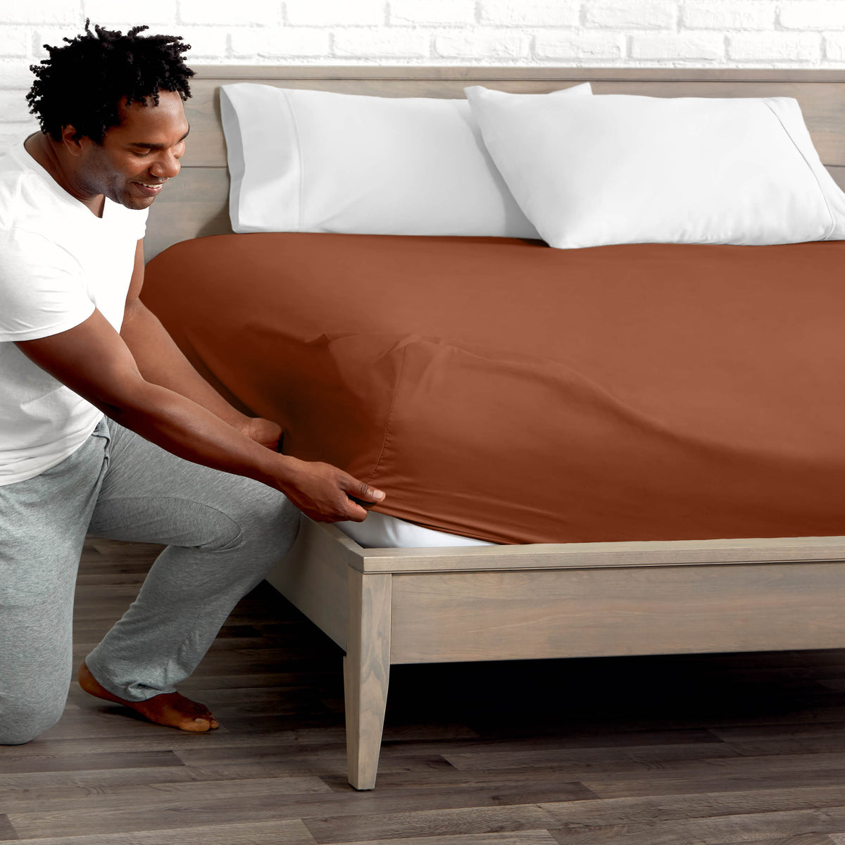 Man adjusting fitted sheet on a bed with white pillows.