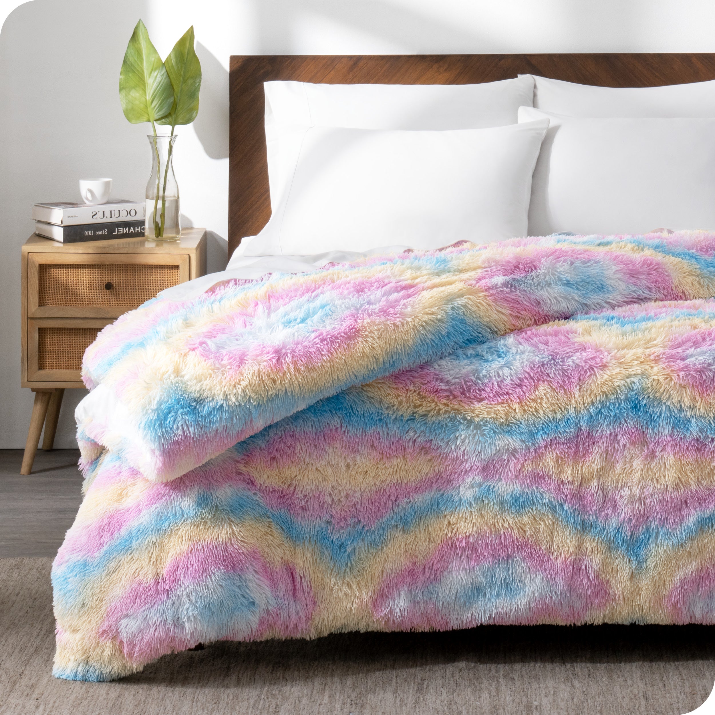 A tie-dye shaggy duvet cover on a bed with white sheets and pillowcases. A large plant is next to the bed.