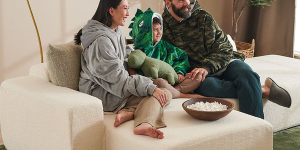 Family sitting on a white sofa in a cozy living room with a plant and lamp in the background.