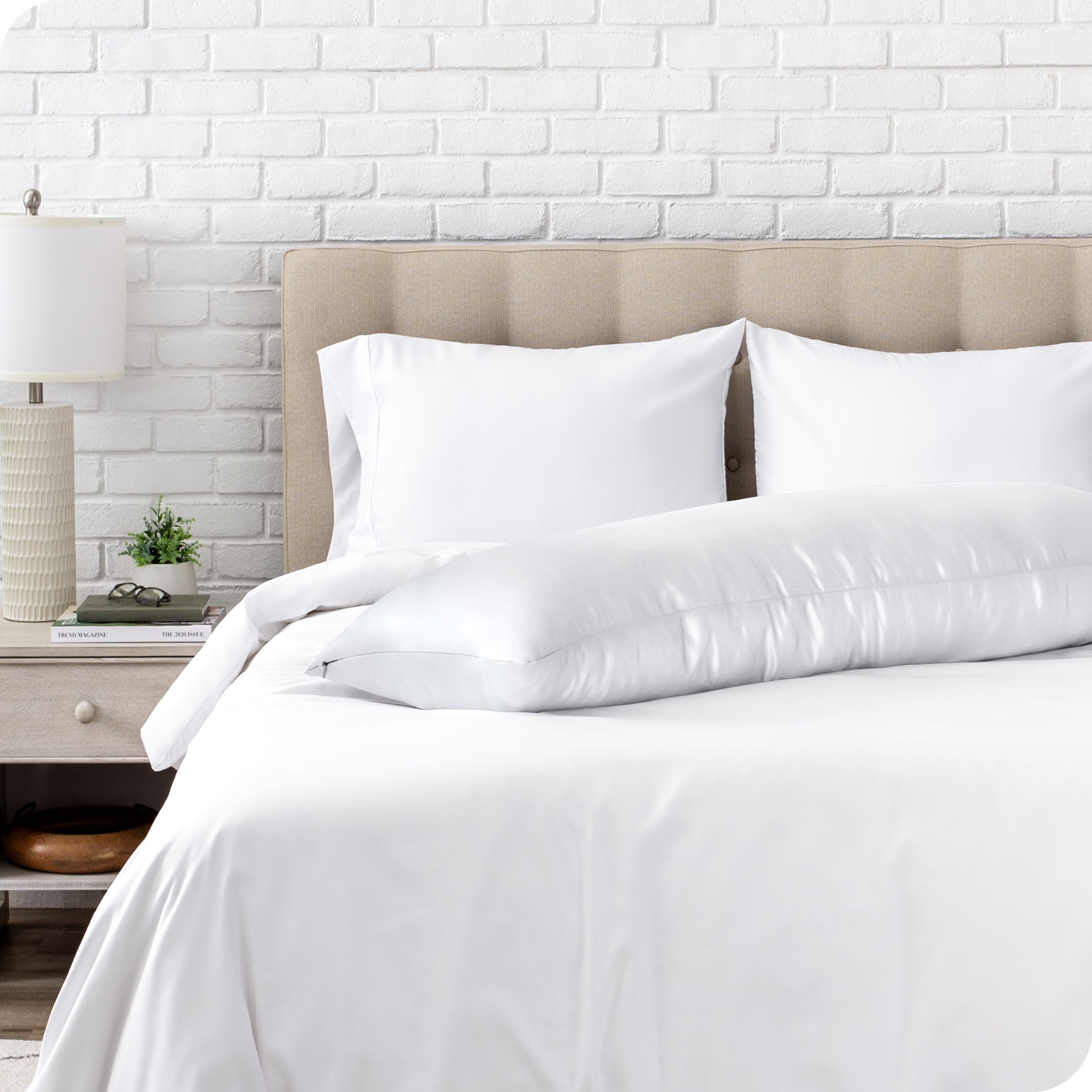Bed with white bedding and a body pillow against a white brick wall.