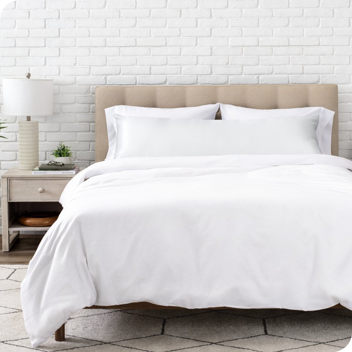 Bed with white bedding and satin pillowcase against a white brick wall.