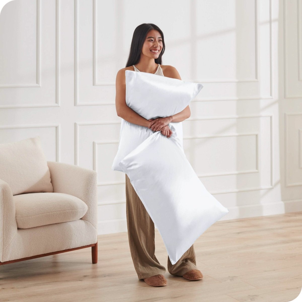A woman is standing and holding 2 pillows covered with silk pillowcases