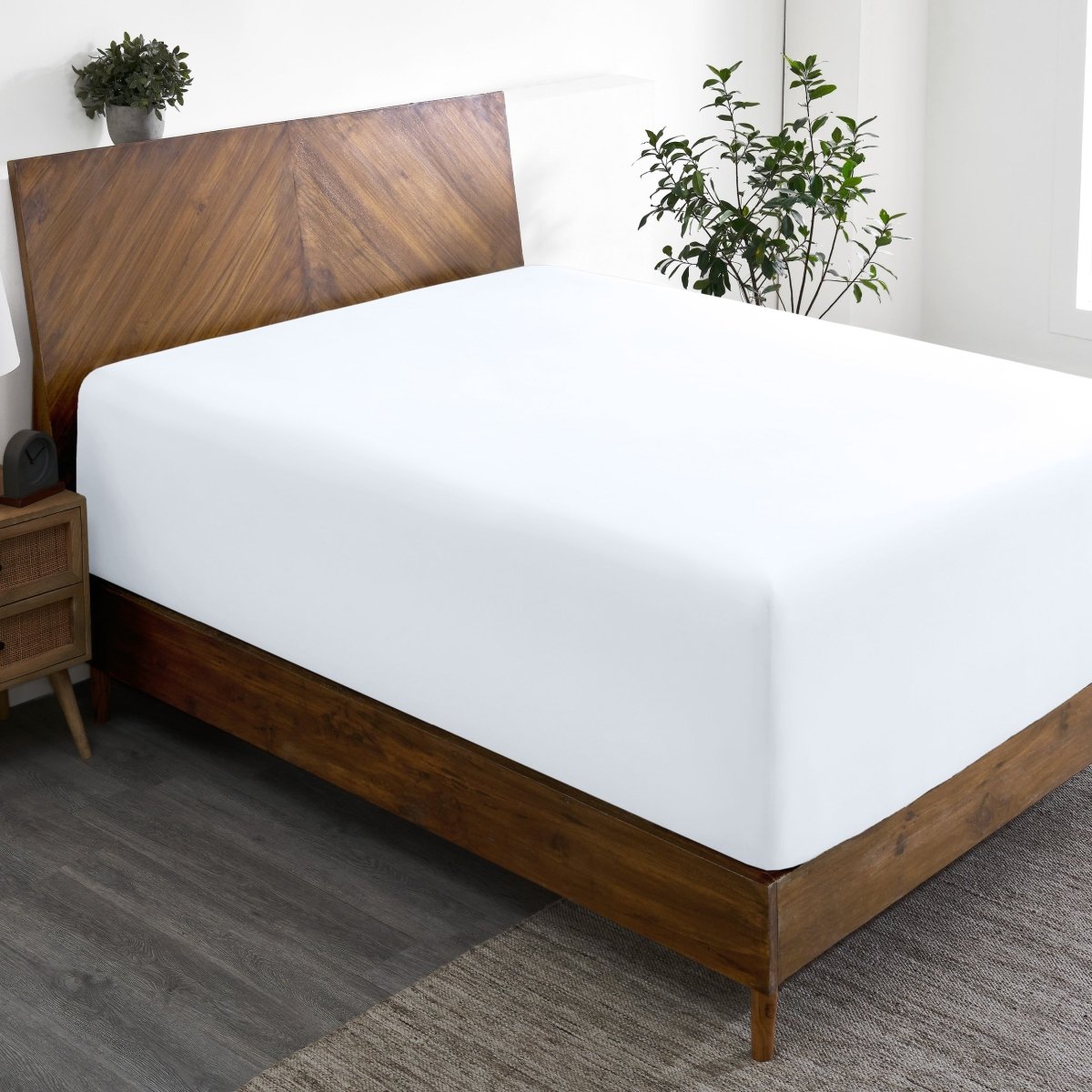 Wooden bed frame with a fitted sheet on the mattress in a room with plants and a nightstand.