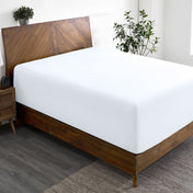 Wooden bed frame with a fitted sheet on the mattress in a room with plants and a nightstand.