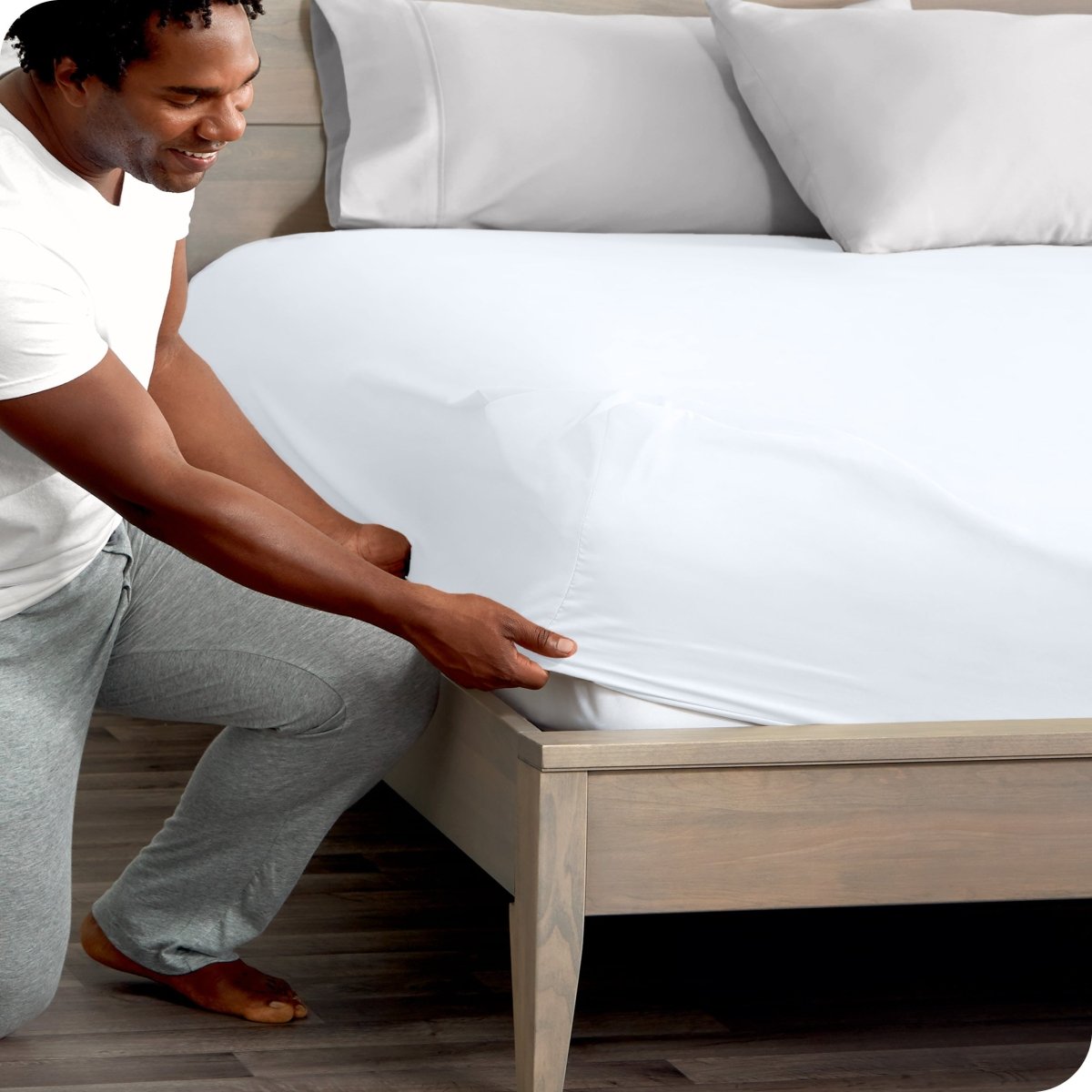 Man in white shirt and gray pants adjusting a fitted sheet on a bed.