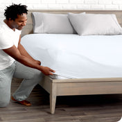 A man is kneeing and putting the corner of the fitted sheet on the mattress. The mattress is on a wooden bed frame.