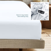 A fitted bed sheet on a bed frame with a small inset close-up of the elastic and a sewn-in tag labeled BOTTOM. White text on the sheet reads, 'Sewn-in tag makes bed-making easy.