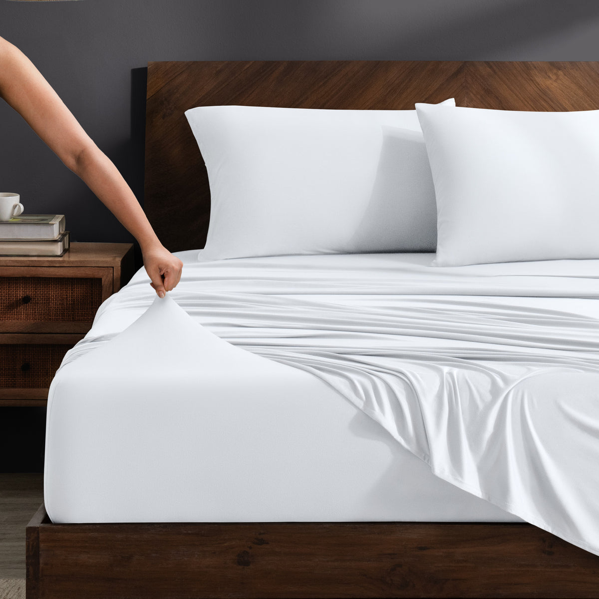 Person adjusting stretch sheets on a bed with a wooden headboard.