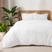 Bed with white duvet set and a wicker headboard, surrounded by potted plants in a bright room.