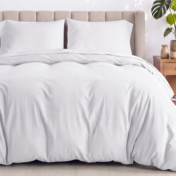 Bed with white duvet set and a wicker headboard, surrounded by potted plants in a bright room.