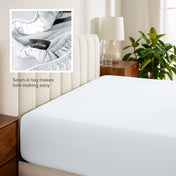Fitted sheet on a bed with a close-up of a sewn-in tag, nightstand, lamp, and plant in a bedroom setting.