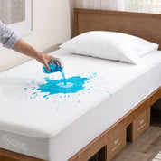 Person pouring liquid onto a white mattress protector in a bedroom setting.