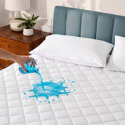 Person pouring blue liquid onto a white mattress protector with a wooden nightstand and plant in the background.