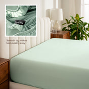 Fitted sheet on a bed with a close-up of a sewn-in tag, nightstand, lamp, and plant in a bedroom setting.