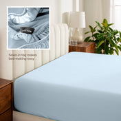 Fitted sheet on a bed with a close-up of a sewn-in tag, nightstand, lamp, and plant in a bedroom setting.