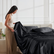 Woman arranging satin sheets on a bed in a bright bedroom.