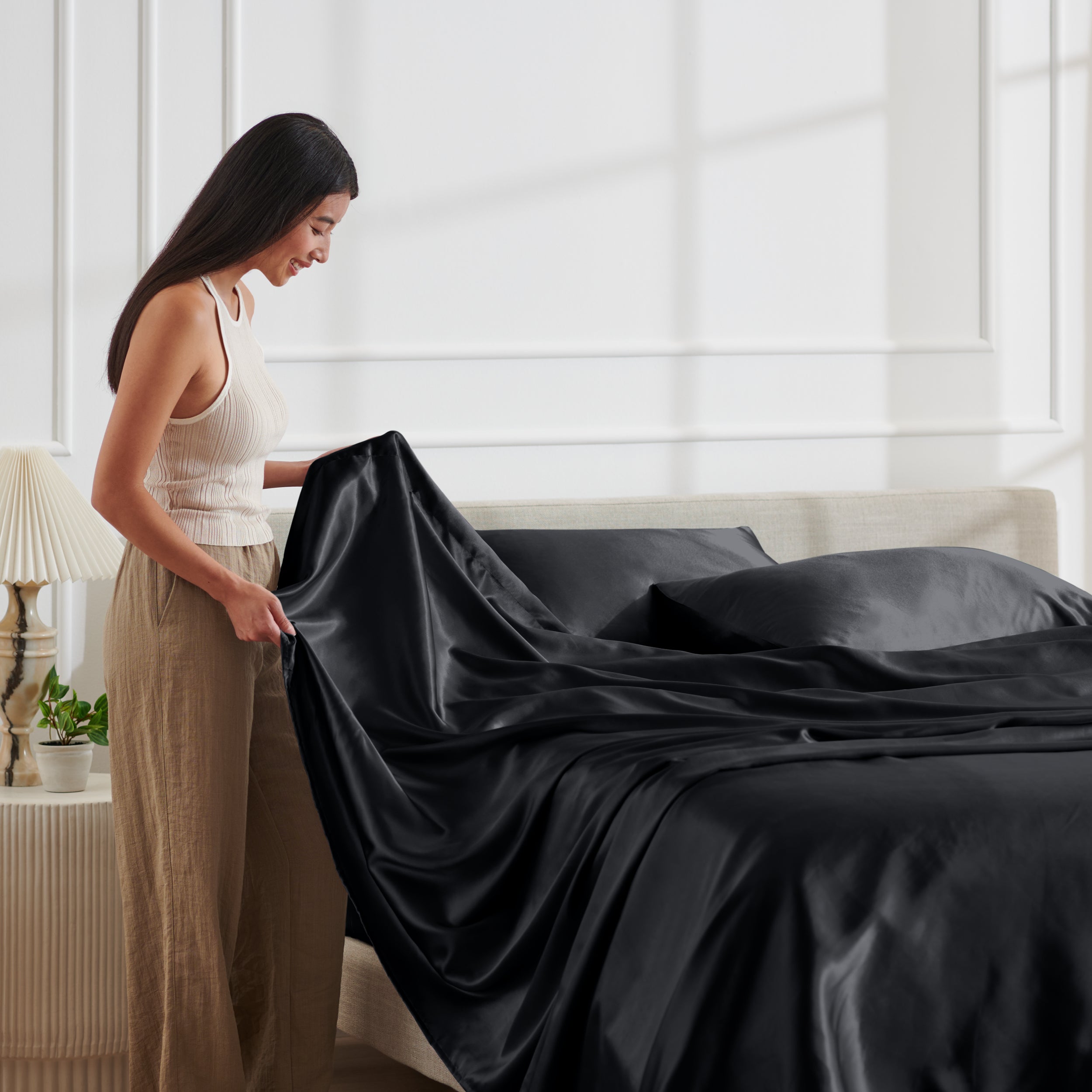 Woman arranging satin sheets on a bed in a bright bedroom.