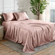 Blush satin bedding set on a bed in a bedroom with a plant and window in the background.