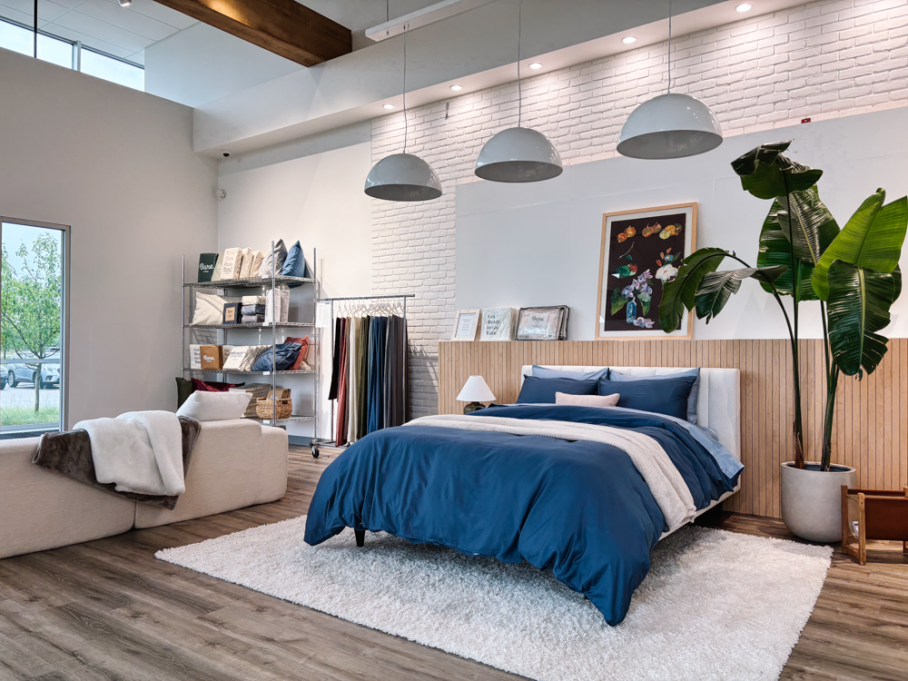 A modern bedroom interior featuring a bed dressed in blue bedding, set on a white rug in a styled room with wood flooring, pendant lights, a white brick wall, and a large potted plant.