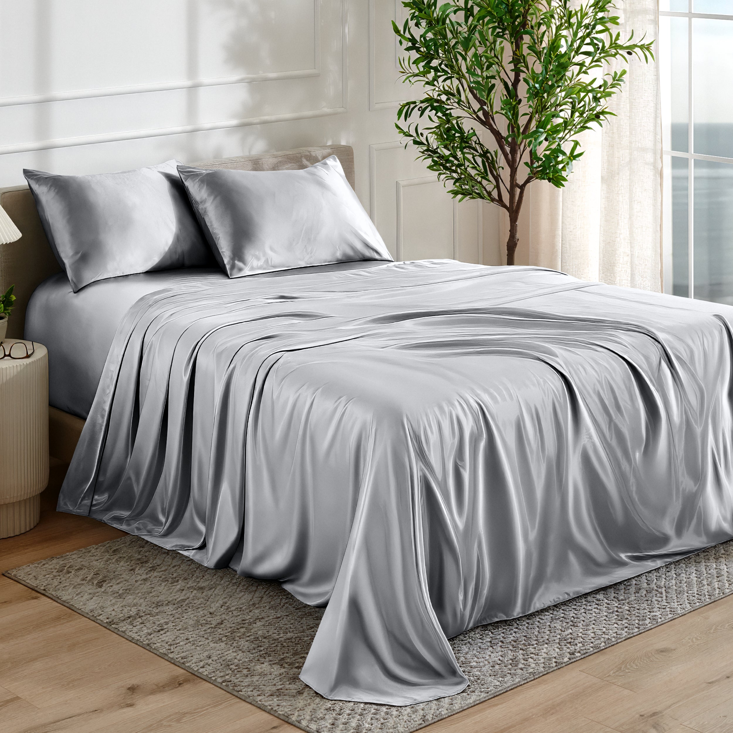 Silver satin bedding set on a bed in a bedroom with a plant and window in the background.
