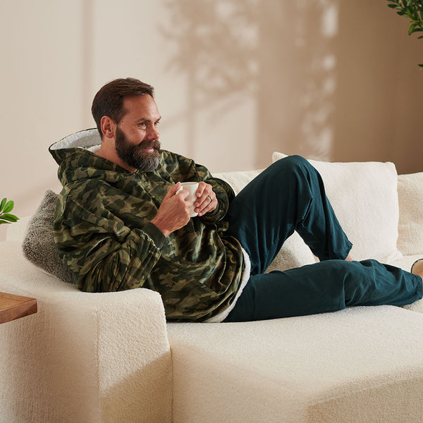Guy in a camo wearable blanket drinking coffee on a white couch.